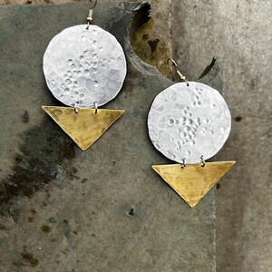 May include: A pair of silver and gold earrings with a hammered texture. The earrings are made of two pieces: a round disc and a triangle. The disc is silver and has a hammered texture. The triangle is gold and has a hammered texture.