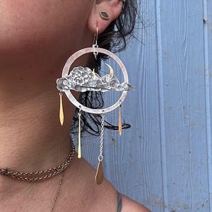 May include: A silver-toned hoop earring featuring a textured cloud shape with a small gold-toned crescent moon. Three gold-toned metal dangles hang from the bottom of the cloud, and a silver-toned chain with a gold-toned teardrop charm hangs below.