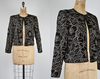 May include: A black velvet jacket with a gold floral embroidery design. The jacket has a  open front and long sleeves. The label on the jacket reads "Collapsible Model 1058".