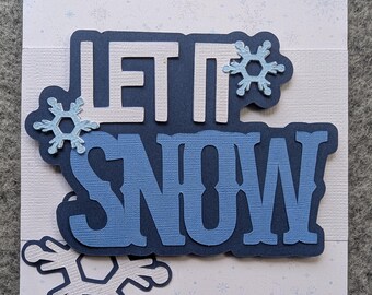 Op de afbeelding: Een wenskaart met de tekst LET IT SNOW in gelaagd marineblauw en lichtblauw karton. Het ontwerp bevat uitgestanste sneeuwvlokaccenten in lichtblauw en wit, tegen een bleke achtergrond met een patroon van kleine, vage blauwe sneeuwvlokjes.