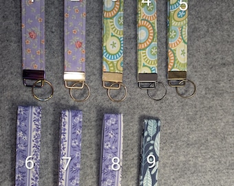 Op de afbeelding: Negen stoffen polssleutelhangers gerangschikt in twee rijen, genummerd van één tot negen. Elk heeft een uniek patroon, waaronder bloemen-, geometrische en botanische prints in tinten paars, groen, blauw en oranje, afgewerkt met metalen hardware.