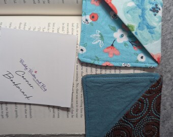 Op de afbeelding: Twee driehoekige stoffen hoekbladwijzers. De ene heeft een lichtblauwe achtergrond met een bloemenpatroon in rood, wit en blauw. De andere is effen gedempt blauw met een donkerbruin driehoekig hoekaccent met een patroon van kleine stippen.