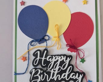 Op de afbeelding: Een handgemaakte wenskaart met een witte, getextureerde achtergrond en een groene rand. Drie papieren ballonnen in blauw, geel en rood zijn bevestigd met bijpassende gekleurde draadjes. De tekst Happy Birthday is in zwart en wit.