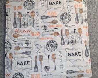 Op de afbeelding: Een vierkante gewatteerde pannenlap in wit met een herhalend patroon van keukengerei, waaronder gardes, lepels en vorken, naast teksten zoals BAKE, BLEND, SALADE en MARKET FRESH in zwarte en oranje inkt met een klein stoffen lusje.