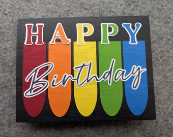 Op de afbeelding: Een rechthoekige wenskaart met een zwarte getextureerde achtergrond met vijf verticale afgeronde strepen in rood, oranje, geel, groen en blauw. Het woord HAPPY is gedrukt in kleurrijke blokletters en Birthday in witte schrijfletters.