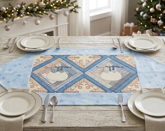 Op de afbeelding: Een rechthoekige tafelloper met een centraal paneel met twee sneeuwpoppen in blauwe en beige ruitvormige kaders, omringd door een lichtblauwe rand met sneeuwvlokpatronen, geplaatst op een houten tafel met witte borden en bestek.