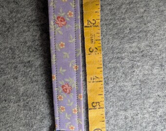 Op de afbeelding: Een lavendelkleurige stoffen polssleutelhanger met een klein bloemenpatroon in rood, geel en groen. De band is bevestigd aan een metalen iriserende sluiting. Een geel meetlint toont een bandlengte van ongeveer 16,5 cm.