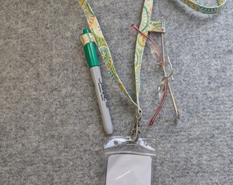 Op de afbeelding: Een stoffen lanyard met een cirkelvormig patroon in groen, geel en blauw met een zwarte plastic gesp. Het bevat een doorzichtige plastic badgehouder, een groene Sharpie-marker in een stoffen lus en een bril met een dun montuur.