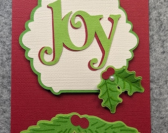Op de afbeelding: Een rechthoekige rode wenskaart met een crèmekleurige, geschulpte uitgestanste vorm met het woord JOY in groene schrijfletters. De kaart is versierd met twee groene hulstblad-uitsparingen, waarvan één met rode bessen en een kleine witte parel.