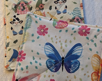 Op de afbeelding: Twee vierkante stoffen onderzetters met doorgestikte naden. De bovenste onderzetter heeft een grote blauwe vlinder, roze bloemen en kleurrijke stippen op een crèmekleurige achtergrond. De onderste onderzetter toont een patroon van kleine kabouters en bloemen.