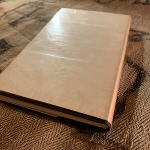 Puede incluir: Un libro de tapa dura de color marr&oacute;n claro con una cubierta lisa y ligeramente texturizada. El libro est&aacute; cerrado y se encuentra sobre una superficie de color marr&oacute;n con un patr&oacute;n.