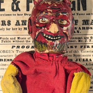 Puede incluir: Una muñeca de diablo vintage con cabeza, cuernos y cuerpo rojos, y guantes amarillos. La cara de la muñeca está pintada con una sonrisa siniestra y ojos negros. El fondo presenta un periódico vintage con las palabras "E & FOREIGN" visibles.