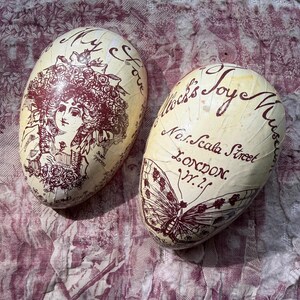 May include: Two cream-coloured, egg-shaped decorative objects with maroon illustrations. One features a woman's portrait and the words "My Love". The other displays a butterfly and text that reads "Mocks Joy Museum, London, W.1."