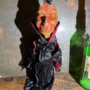 Puede incluir: Una figura de diablo con rostro naranja texturizado, ojos amarillos y dos cuernos grises. El personaje viste una capa de satén negro con forro rojo oscuro, que presenta un cuello alto y rígido que enmarca el rostro y tela drapeada.