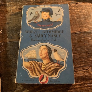 Puede incluir: Una cubierta de libro azul con una imagen de un espantapájaros con un sombrero azul y un pájaro en una rama. El título es "Worzel Gummidge & Saucy Nancy" de Barbara Euphan Todd. Es un libro de la colección Puffin Story Book.