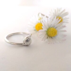 May include: A silver ring with a textured, round, silver band. The ring is on a white surface with white daisies in the background.