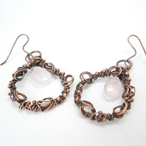 May include: A pair of copper wire earrings with a pink rose quartz stone hanging from each. The earrings are made of intricate wirework and have a delicate, feminine look.