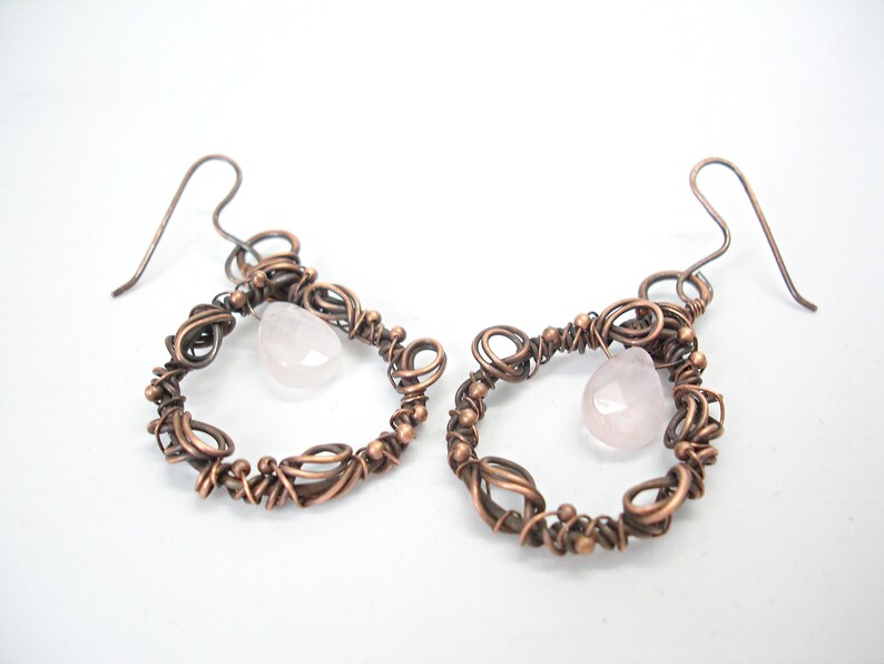 May include: A pair of copper wire earrings with a pink rose quartz stone hanging from each. The earrings are made of intricate wirework and have a delicate, feminine look.