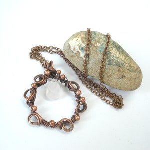 May include: A copper wire wrapped pendant necklace with a clear quartz crystal. The necklace is on a brown rock and a copper chain.