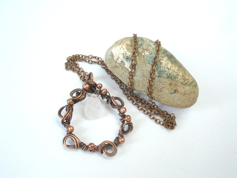 May include: A copper wire wrapped pendant necklace with a clear quartz crystal. The necklace is on a brown rock and a copper chain.