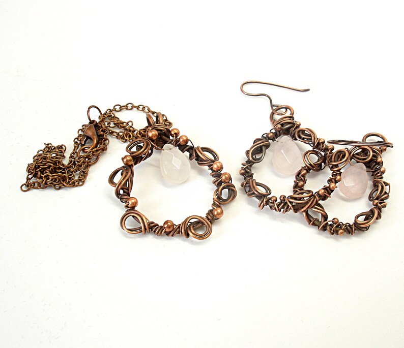 May include: A set of copper wire earrings and a necklace with rose quartz stones. The earrings are made of three intertwined circles with a rose quartz stone hanging from each circle. The necklace is a single circle with a rose quartz stone hanging from the bottom.