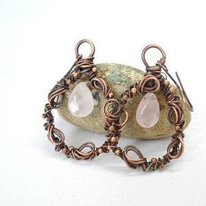 May include: A pair of copper wire earrings with rose quartz drops. The earrings are made of twisted copper wire and have a delicate, intricate design.