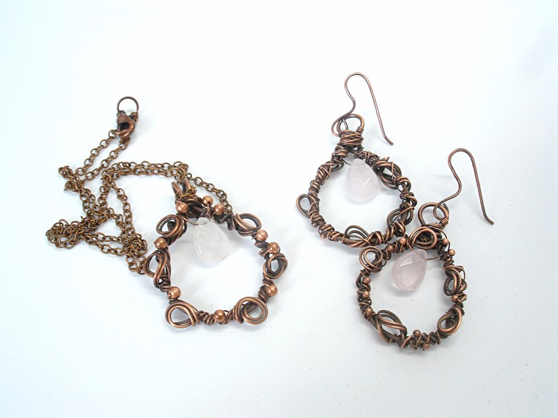 May include: A set of copper wire earrings and a necklace with pink rose quartz stones. The earrings and necklace feature a delicate, intricate design.