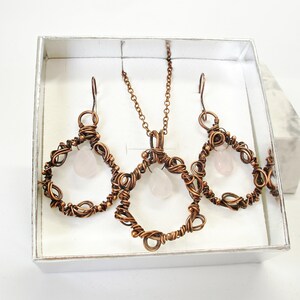 May include: A copper wire jewelry set with a rose quartz pendant and earrings. The pendant and earrings are in the shape of a circle with a rose quartz stone in the center.