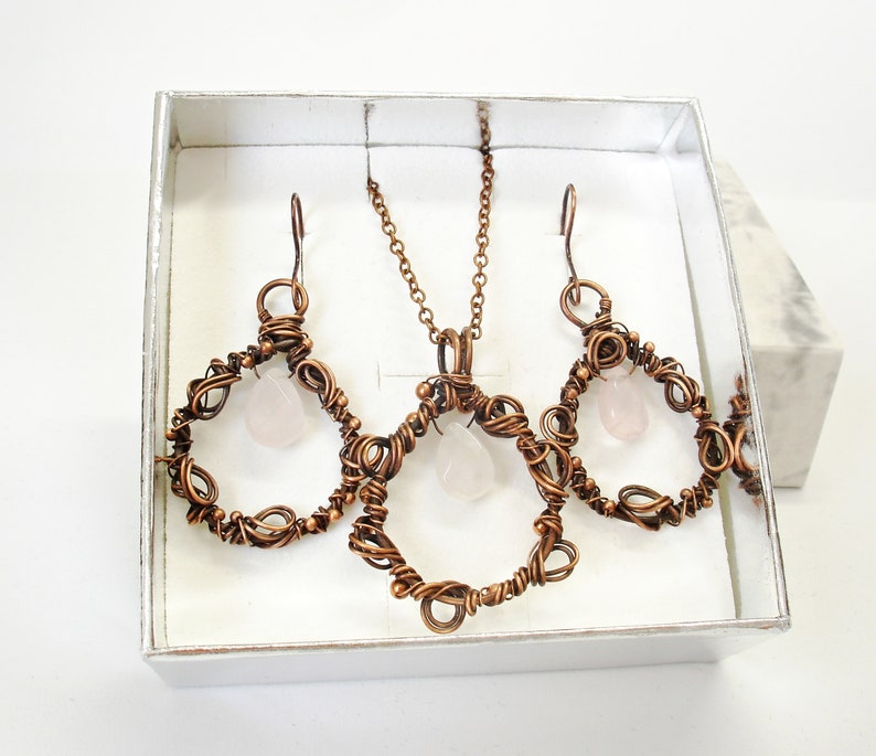 May include: A copper wire jewelry set with a rose quartz pendant and earrings. The pendant and earrings are in the shape of a circle with a rose quartz stone in the center.