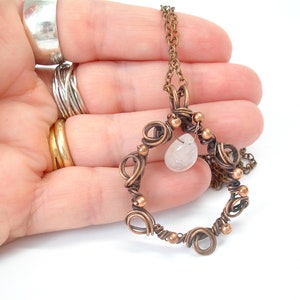 May include: A copper wire wrapped pendant necklace with a clear quartz stone. The pendant is in the shape of a circle with copper wire wrapped around it. The necklace is on a copper chain.