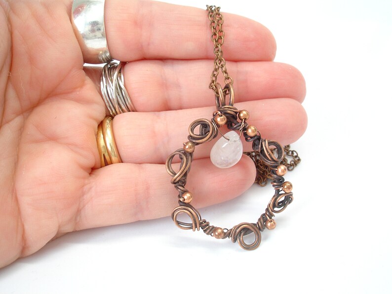 May include: A copper wire wrapped pendant necklace with a clear quartz stone. The pendant is in the shape of a circle with copper wire wrapped around it. The necklace is on a copper chain.