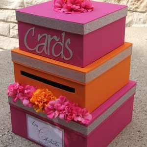 May include: A three-tiered card box with a pink, orange, and pink color scheme. The top tier has a silver glitter trim and the words "Cards" in silver lettering. The bottom tier has a silver glitter trim and a label with the text "Leo & Astrid" and the date "August 22, 2014". The box is decorated with pink and orange flowers.