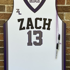 May include: White basketball jersey with purple trim and a black number 13. The jersey has the name "ZACH" printed in black letters and the date "09.04.21" printed in black letters on the neckline. Two purple markers are hanging from the jersey.
