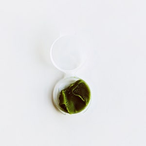May include: A small, round, clear plastic container with a green, creamy substance inside.