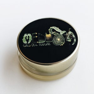 May include: A small, round, gold-colored tin with a black lid. The lid has a green and gold design with the words "MEDUSA WILD OIL PERFUME".