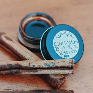 May include: A small glass jar with a black lid and a blue label that reads "Wild Veil Cinnamon Bark Absolute". The jar is sitting on a wooden surface with cinnamon sticks.