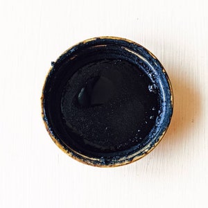 May include: A small, round, brown container with a black liquid inside. The container has a slightly raised rim.