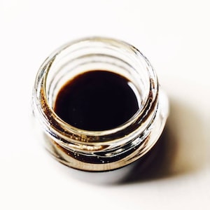 May include: A small glass jar filled with a dark brown liquid. The jar has a clear glass lid.