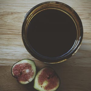 May include: Overhead shot of a glass jar filled with a dark liquid, possibly balsamic vinegar or a similar condiment. Two halves of a fresh fig, showing the red interior, are placed beside the jar on a wooden surface.