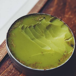 May include: A small round tin container filled with a green balm or ointment. The balm has a textured surface and appears to be made with natural ingredients.