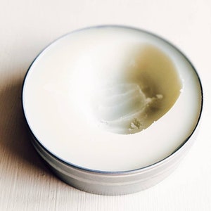 May include: A white cream in a silver tin container. The cream is partially used, leaving a dip in the centre.