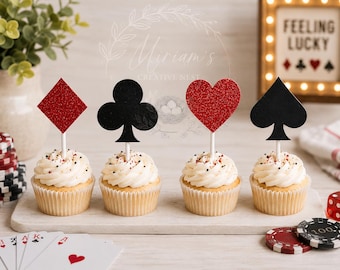 May include: Four cupcakes topped with white frosting and card suit toppers. The toppers feature a red glitter diamond, a black clover, a red glitter heart, and a black spade, each attached to a white stick and inserted into the center of the cupcake.