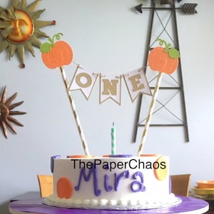 May include: A white cake with purple and orange frosting decorated with a pumpkin themed cake topper. The topper includes a banner that says "ONE" in gold glitter letters and two orange pumpkins with green stems. The cake also has the name "Mira" written in purple frosting and the words "The Paper Chaos" written in black frosting.