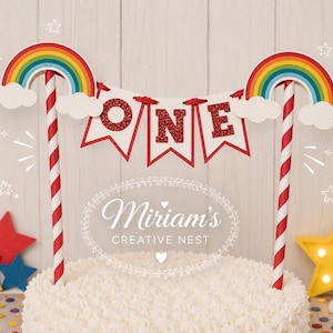 May include: A cake topper featuring two red and white striped paper straws supporting a banner with the word ONE in red glitter letters. Two rainbow arches with white clouds sit atop the straws. The topper is placed on a white frosted cake.
