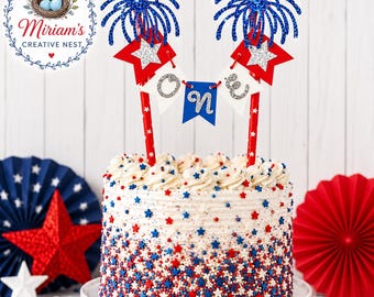 May include: A cake topper featuring two red striped straws topped with blue glittery firework shapes and red star cutouts. Between the straws, three small white and blue pennants spell out the word ONE in silver glittery letters.