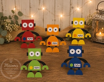 May include: Five colorful paper robot cutouts in red, orange, yellow, green, and blue. Each robot features a square head with two circular eyes, a semi-circular body with a small circular button, a jagged mouth, and black arms and legs.