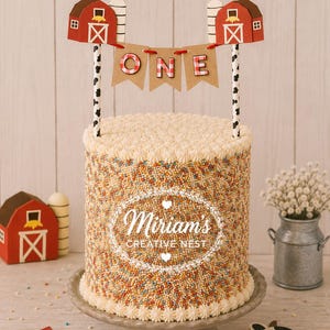 May include: A cylindrical cake covered in multicolored sprinkles with a white piped border. A cake topper features two red barns connected by a brown banner with the word ONE in red and white checkered letters, supported by black and white cow-print sticks.