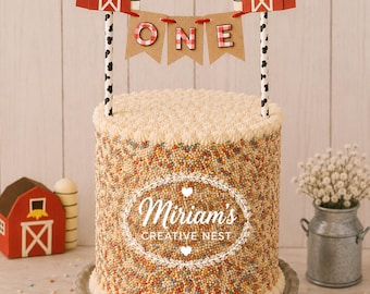 May include: A cylindrical cake covered in multicolored sprinkles with a white piped border. A cake topper features two red barns connected by a brown banner with the word ONE in red and white checkered letters, supported by black and white cow-print sticks.
