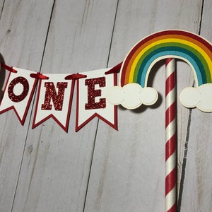 May include: Red glitter cake topper with the word "ONE" on a white banner with red trim. The topper also includes a red and white striped straw with a rainbow and clouds on top.
