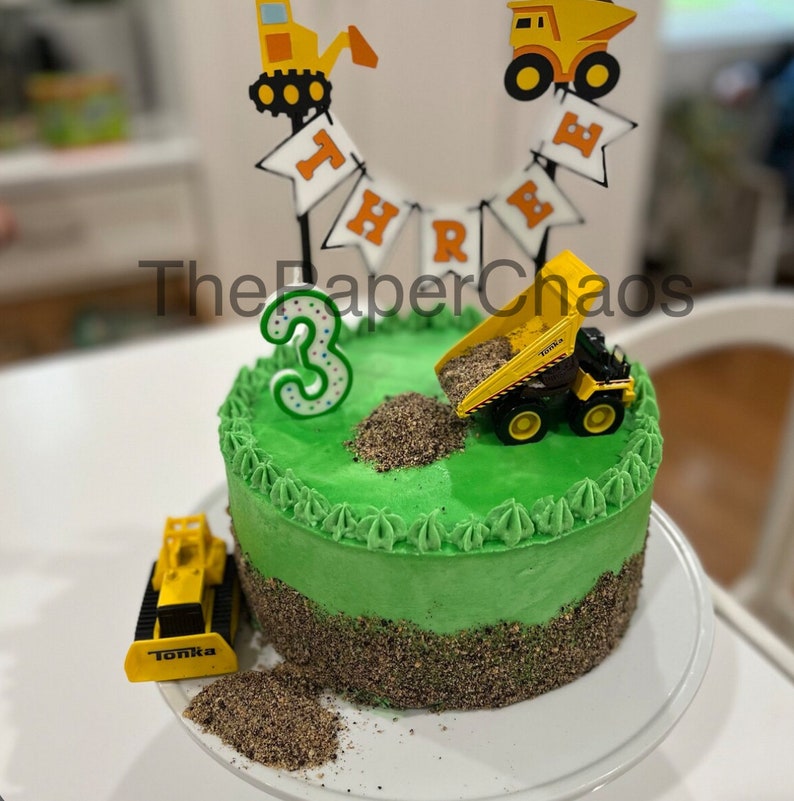 May include: A round cake with green frosting and a brown textured base. It features a green number three candle, a yellow dump truck toy, a yellow construction vehicle toy, and a banner topper with the text THREE and construction vehicle icons.
